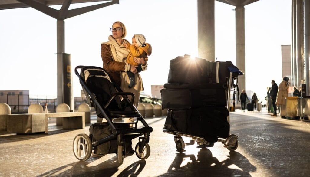 Mother-at-travelling-with-his-infant-baby-boy-child-walking-pushing-baby-stroller-and-luggage-cart-in-front-of-airport-terminal-station